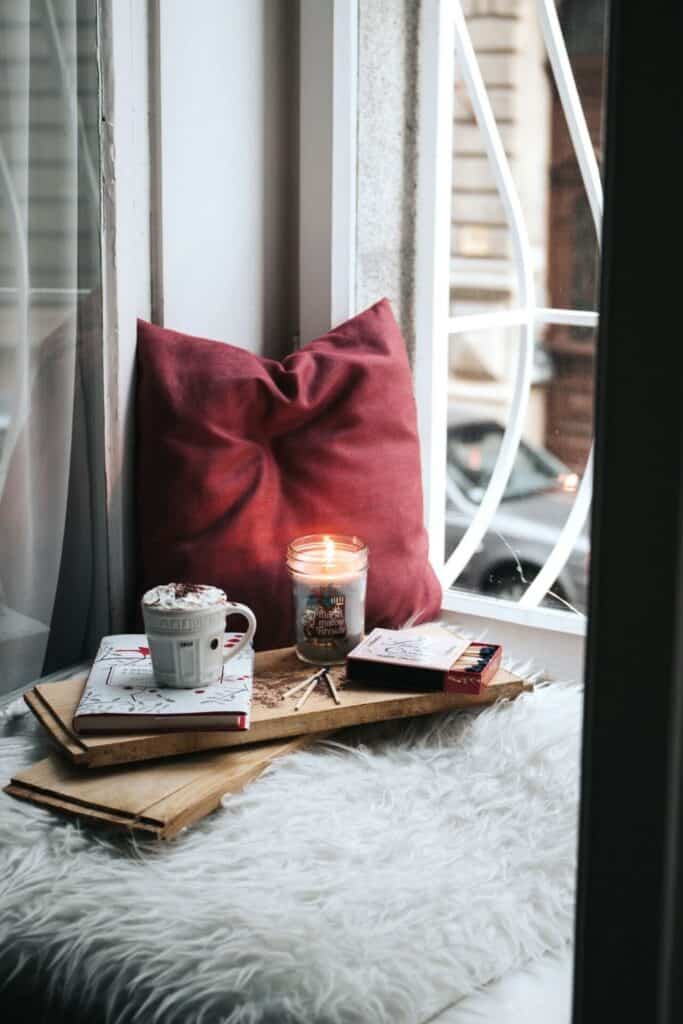 a window seat with a burning candle books, hot chocolate and matches overlooking a calm city street